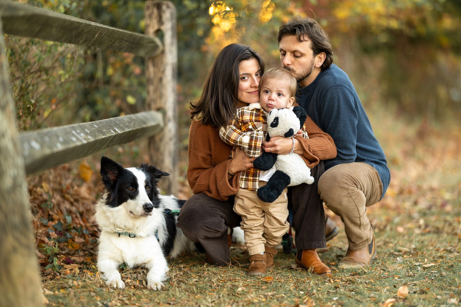 Fall family photo of parents, toddler, and dog next to wood fence