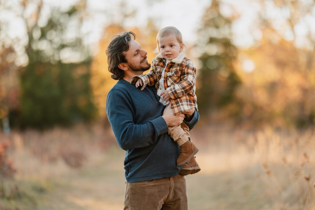Fall family photo of father holding toddler son in front of foliage